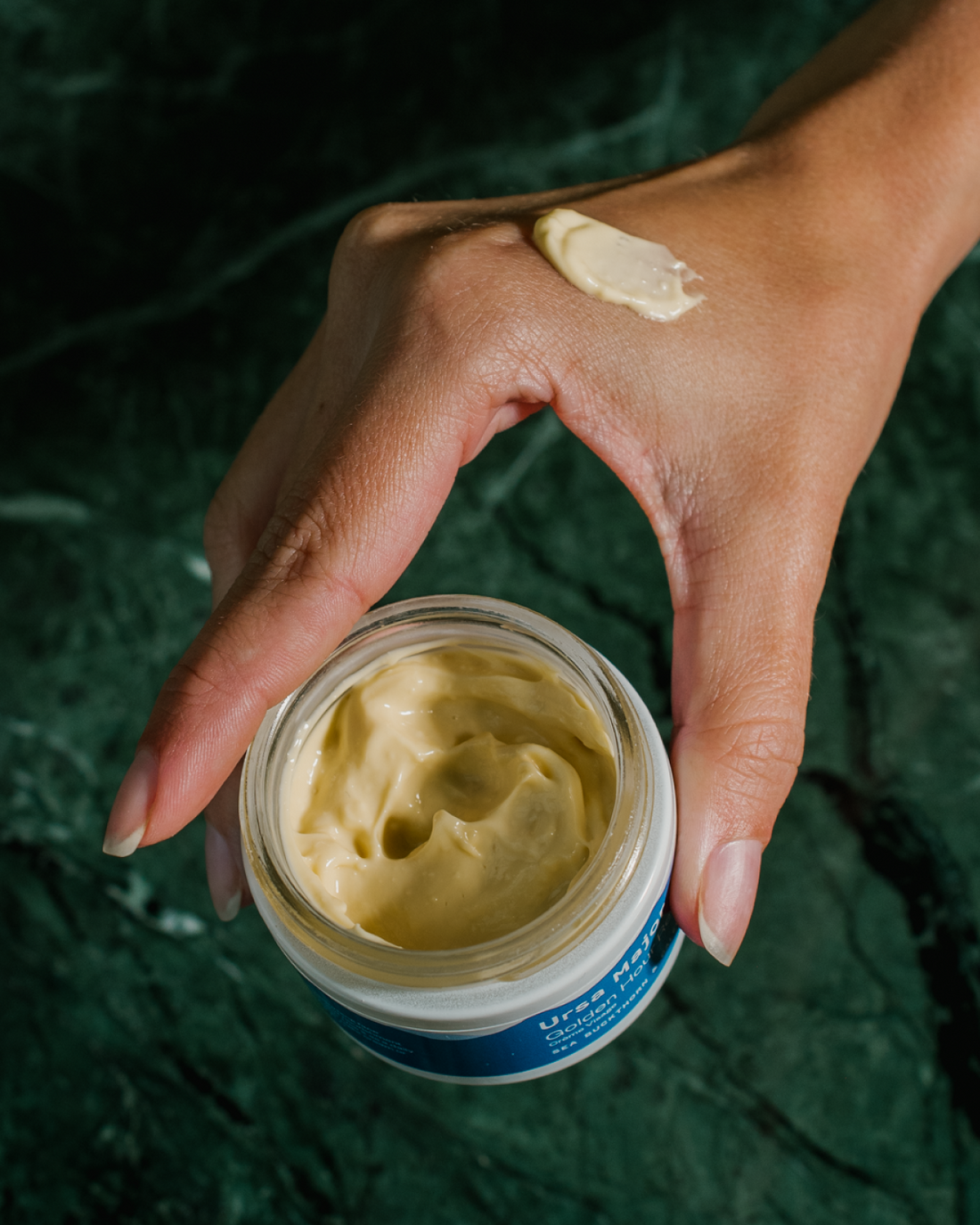 Holding an open Golden Hour Recovery Cream with a smear on hand.