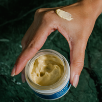 Holding an open Golden Hour Recovery Cream with a smear on hand.